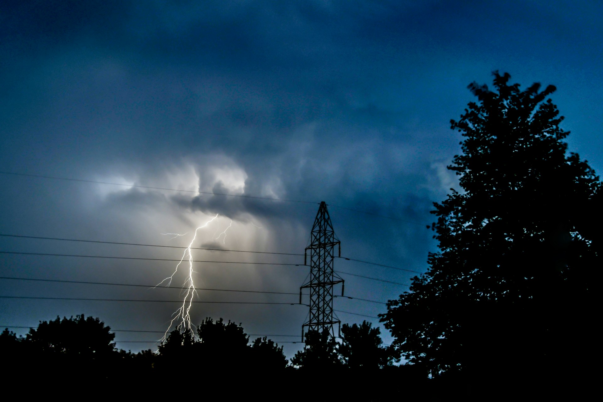 A lightning strike with an electrical transmission tower in the foreground surrounded by trees