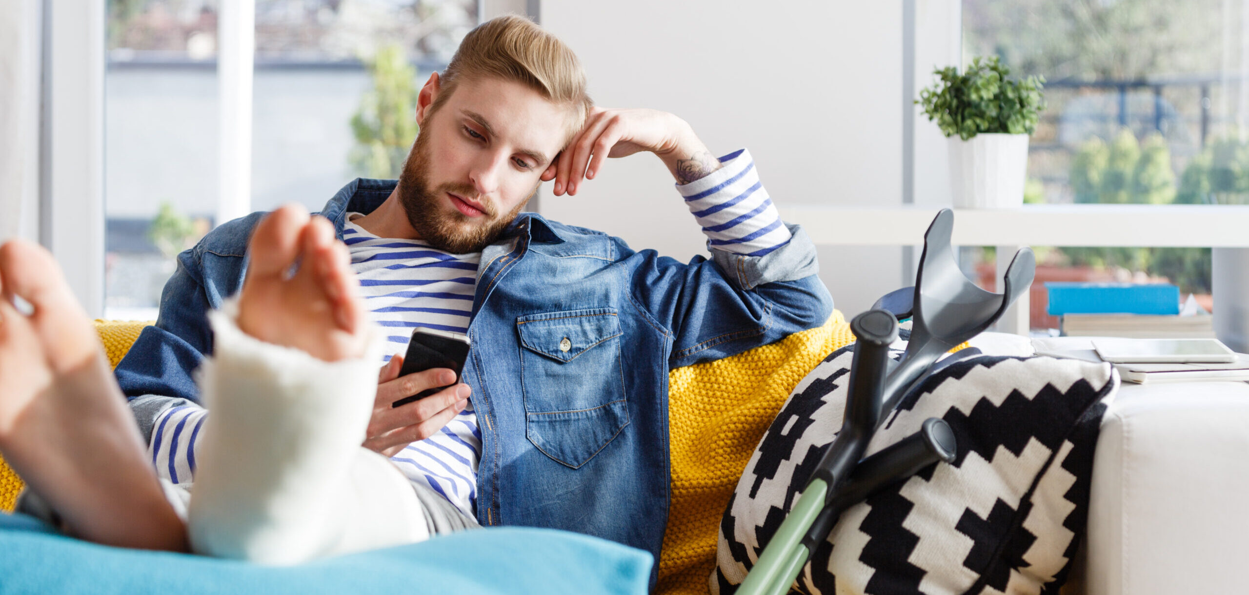 Young man with broken leg in plaster cast lying down on sofa at home and using a smart phone.