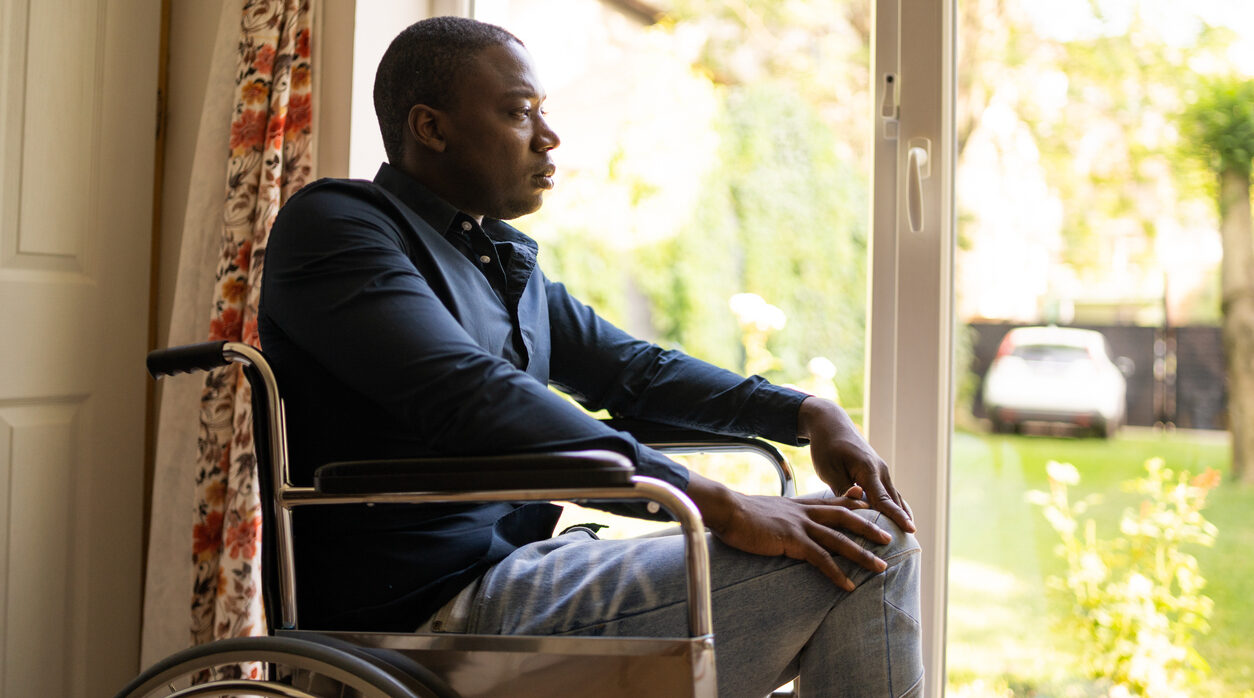 A man is staring from his seat in a wheelchair towards a bright sunny day out a window
