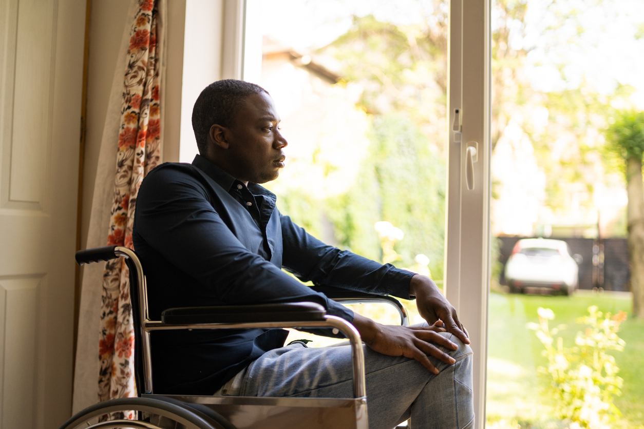 A man is staring from his seat in a wheelchair towards a bright sunny day out a window